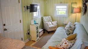 A room with a green-striped wall, a white door, a television mounted above a cabinet, a patterned armchair, a sofa with pillows, and a lamp on a side table. - Escape to this charming 1-bedroom Cape cottage, .4 miles to the beach (Dennis Port)