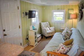 Ein Zimmer mit einer grün gestreiften Wand, einer weißen Tür, einem über einem Schrank montierten Fernseher, einem gemusterten Sessel, einer Couch mit Kissen und einer Lampe auf einem Beistelltisch.