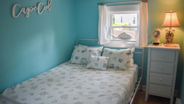 A bedroom with a bed, a nightstand, a lamp, a window with curtains, and a wall-mounted sign. - Escape to this charming 1-bedroom Cape cottage, .4 miles to the beach (Dennis Port)