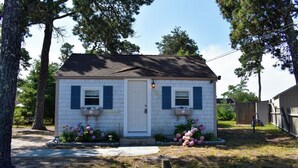 A small, single-story building with a white exterior and blue shutters, surrounded by a wooden fence and trees. - Escape to this charming 1-bedroom Cape cottage, .4 miles to the beach (Dennis Port)