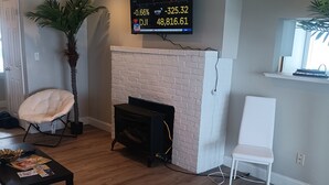 Una sala de estar con chimenea, un televisor mostrando datos del mercado de valores, una silla blanca y un sofá.
