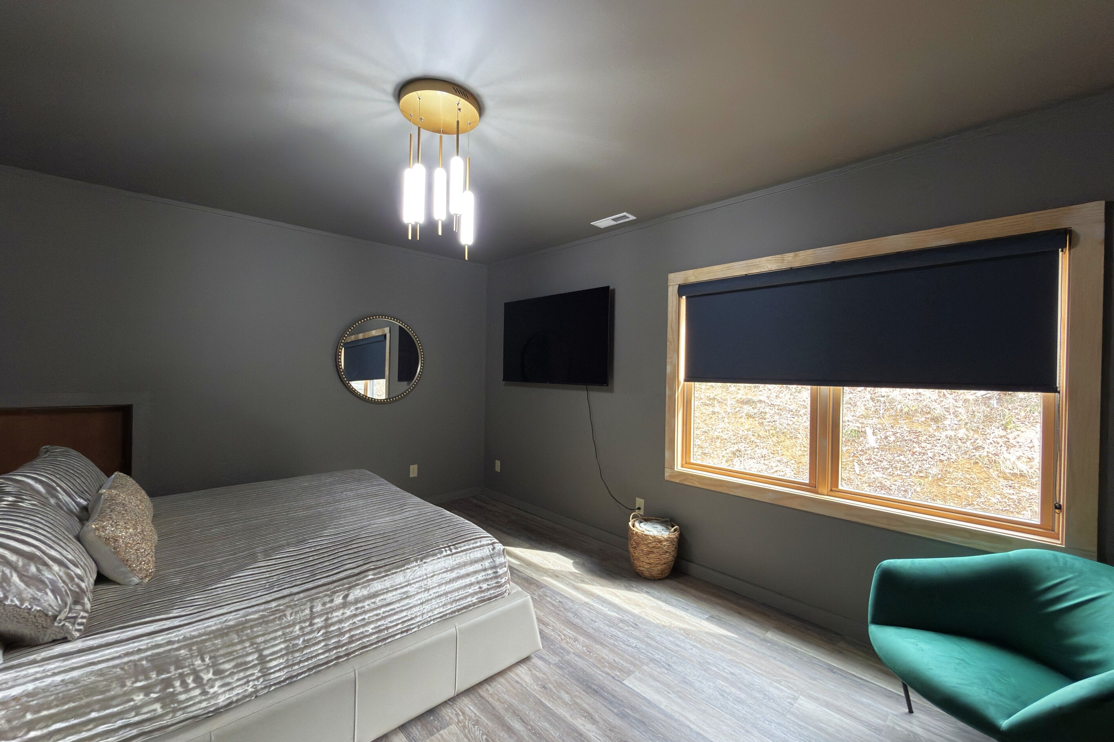 Quarto com uma cama, uma cadeira verde, uma televisão, uma janela com persiana e um lustre.