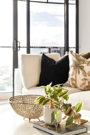 Ein Zimmer mit einem weißen Sofa, einem großen Fenster, einem gewebten Korb, einer Topfpflanze und einem Buch.