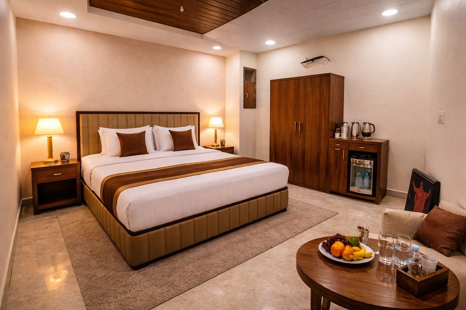 Quarto de hotel com uma cama grande, cabeceira de madeira, duas luminárias de cabeceira, um armário, frigobar e uma pequena mesa redonda com frutas e copos.