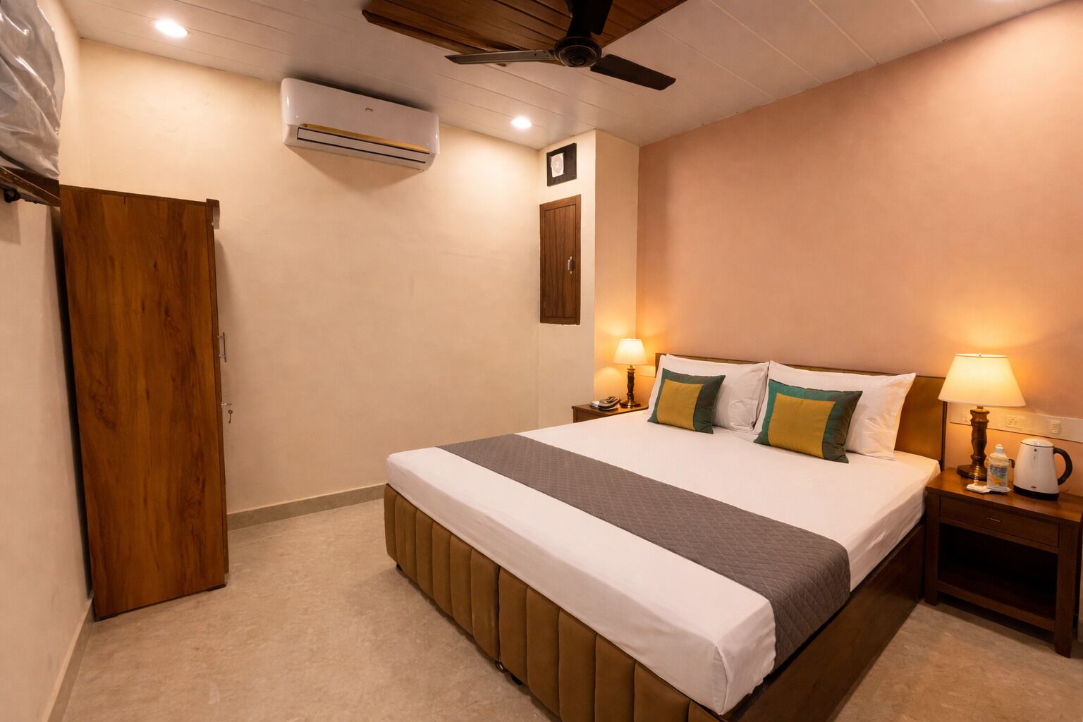 A hotel room with a bed, bedside tables, a wooden door, an air conditioning unit, and a ceiling fan.