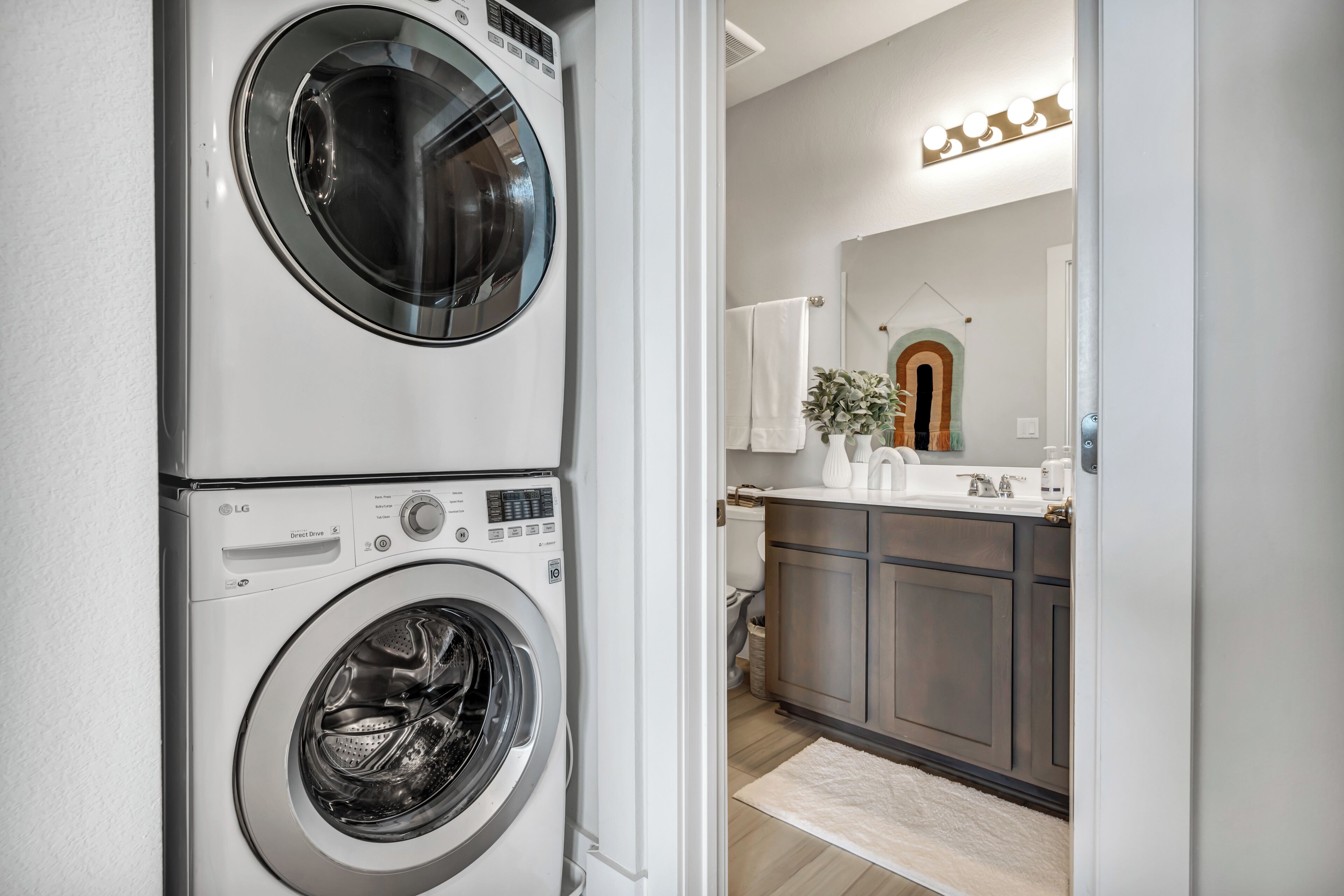 A laundry room with stacked washers and dryers, a kitchen with dark cabinets, and a mirror with lights above the sink.