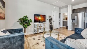 Een moderne woonkamer met een flatscreen televisie, twee blauwe banken, een glazen salontafel, een groot abstract schilderij, een potplant en een keuken met een roestvrijstalen koelkast.