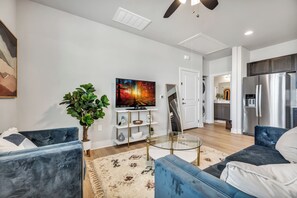 Un salon moderne avec un téléviseur à écran plat, deux canapés bleus, une table basse en verre, un grand tableau abstrait, une plante en pot et un coin cuisine avec un réfrigérateur en acier inoxydable.