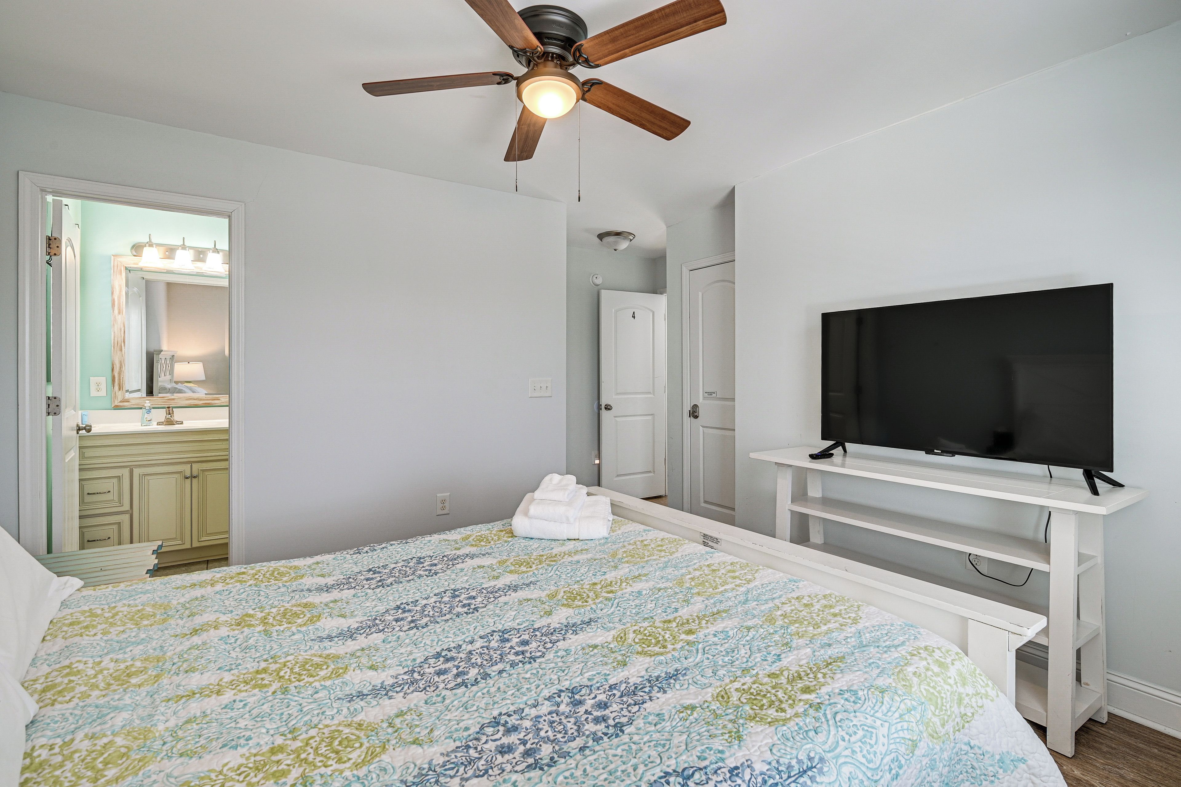 A bedroom with a bed, a ceiling fan, a TV on a stand, and a bathroom visible through an open door.