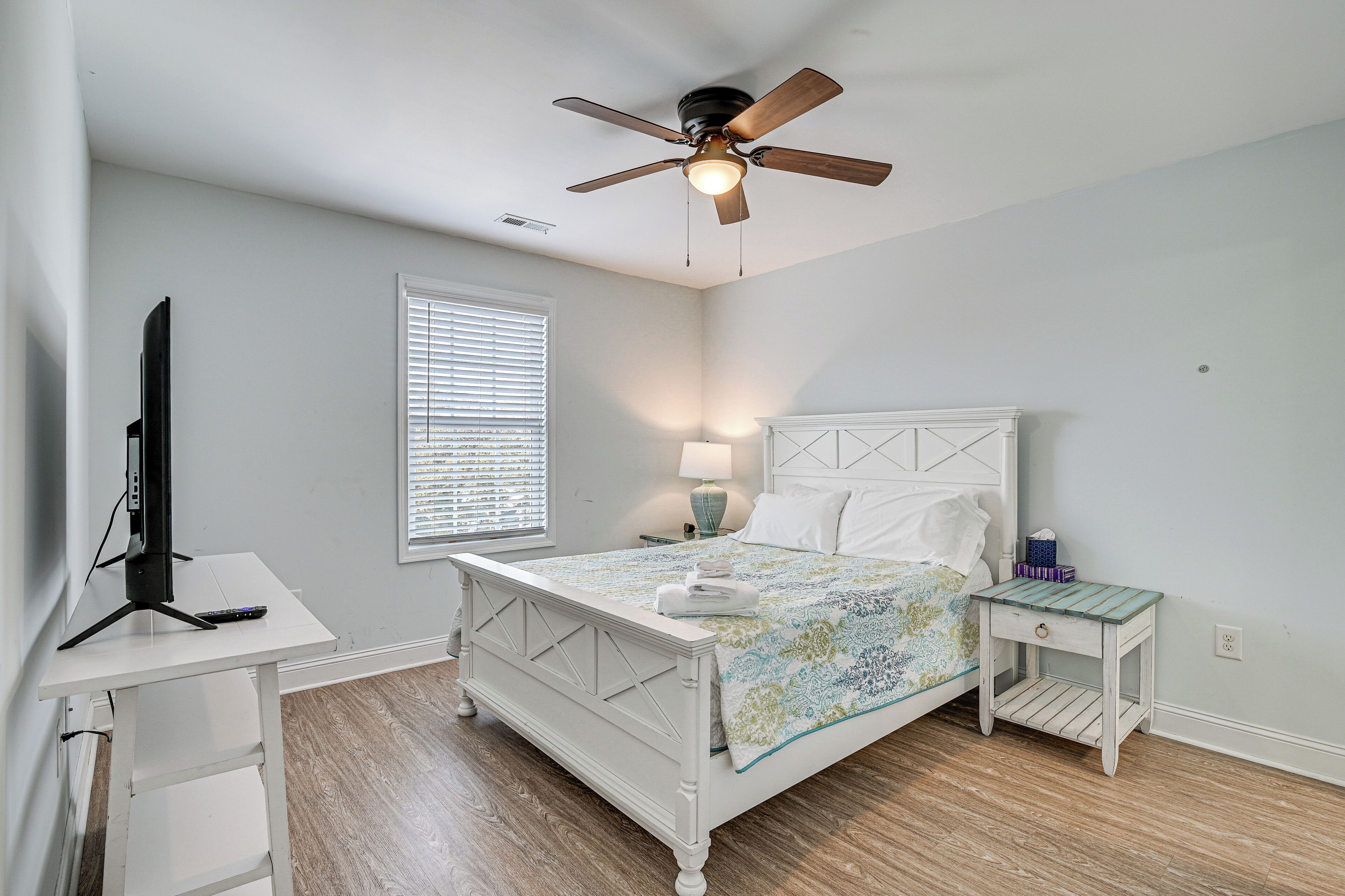A bedroom with a bed, a desk, a ceiling fan, a window with blinds, and a nightstand with a lamp.