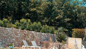 A swimming pool with lounge chairs, a stone wall, and a forested background. - Villa Maluan (Mons)