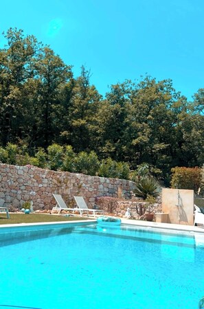Ein Swimmingpool mit Liegestühlen, einer Steinmauer und bewaldeter Landschaft im Hintergrund.