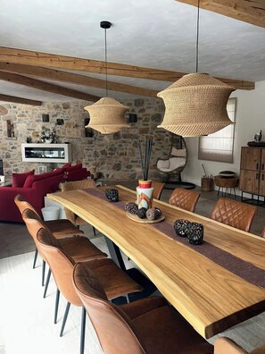 Ein Essbereich mit einem langen Holztisch, braunen Ledersesseln, gewobenen Pendelleuchten und einer Wand aus Stein.