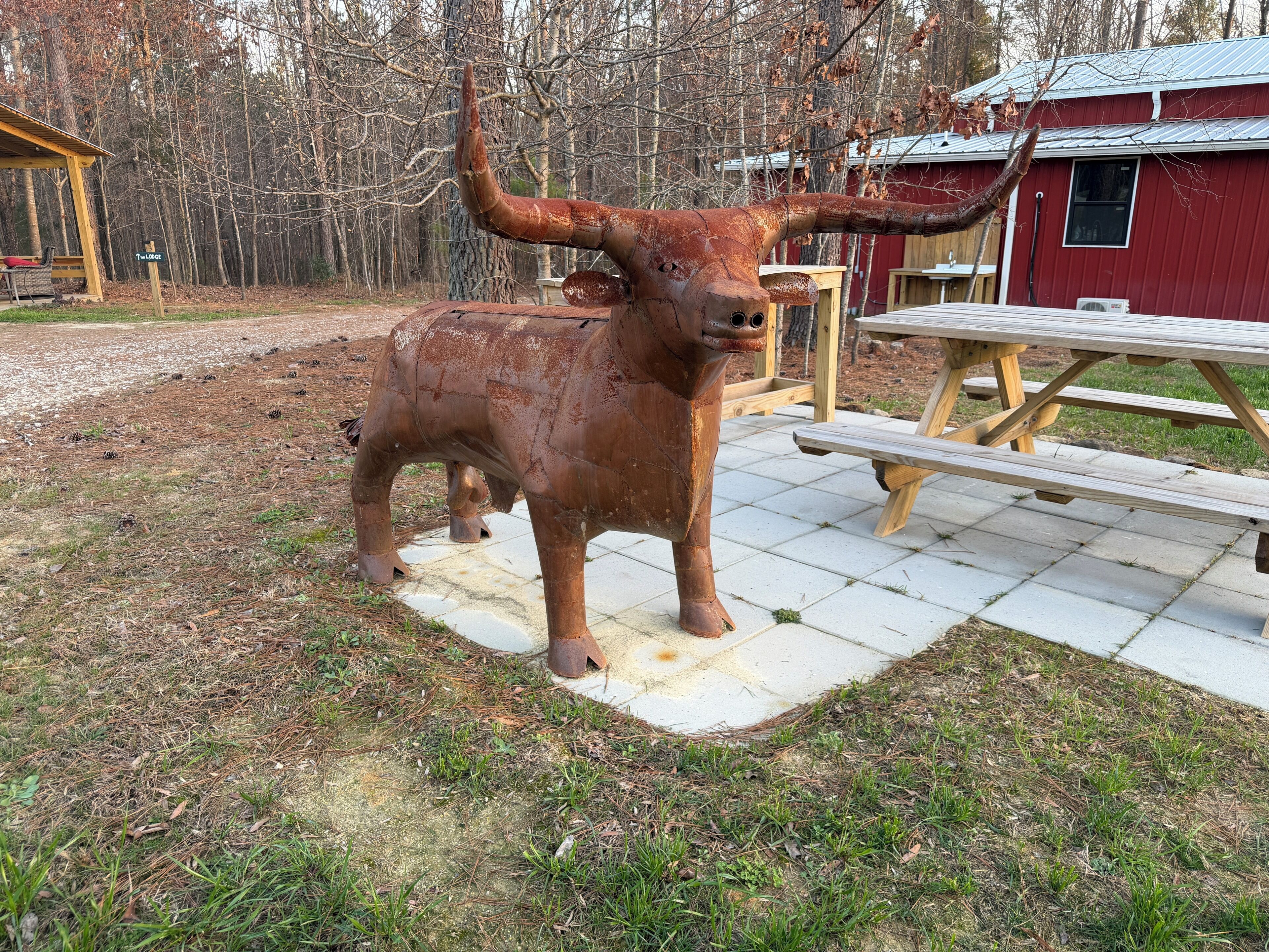 A rustic outdoor area with a large metal bull sculpture, a picnic table, and a red building.