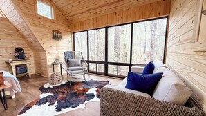 Ein gemütliches Blockhaus mit großem Fenster, Holzwänden, einem gewebten Sessel, einer Couch mit Kissen und einem Kuhfellteppich.
