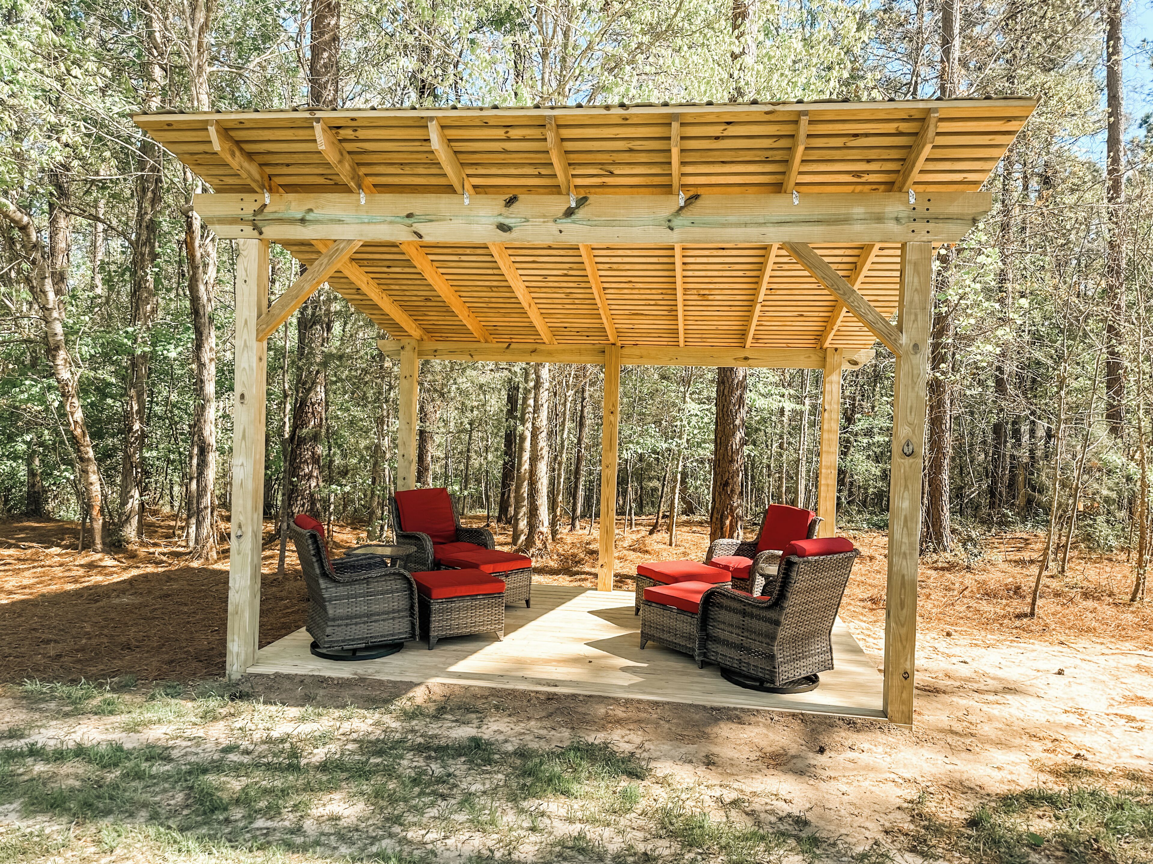 A wooden pergola with wicker chairs and red cushions in a forest setting.