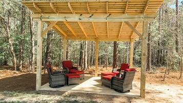 A wooden pergola with wicker chairs and red cushions in a forest setting.
