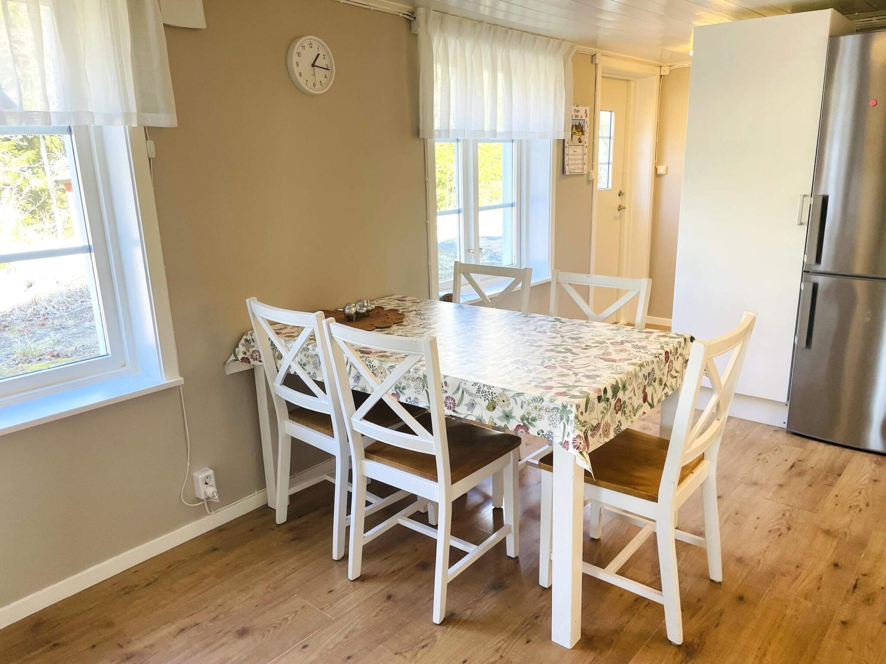 A dining area with a table and chairs, a clock on the wall, and a refrigerator in the background.