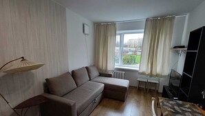 Ein Wohnzimmer mit einer Eckcouch, einem kleinen Tisch, einem Fernseher und einem Fenster mit Vorhängen.