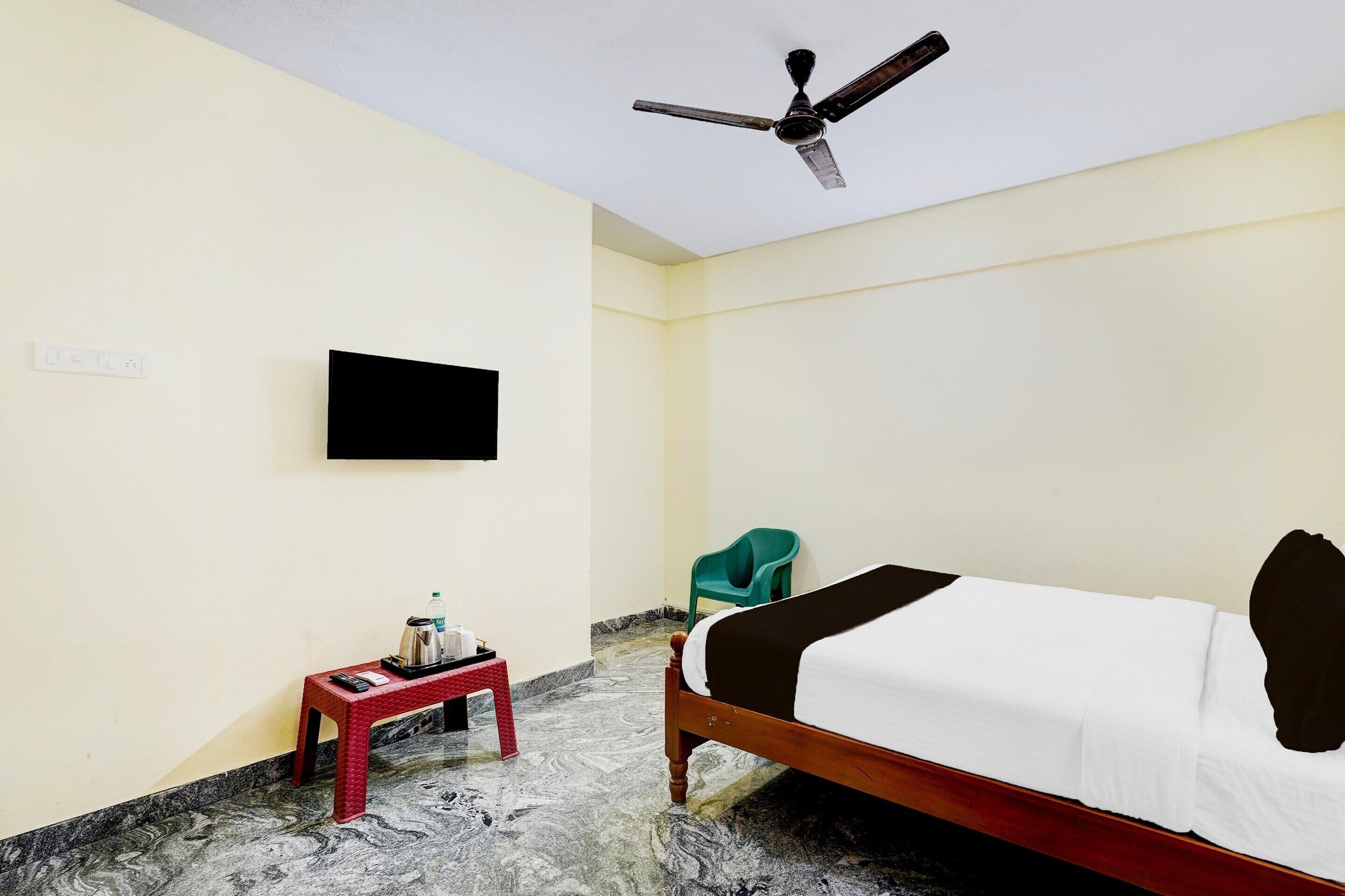 Une chambre d’hôtel avec un lit, un ventilateur de plafond, un téléviseur fixé au mur, une petite table avec une bouteille d’eau et un verre, et une chaise verte.