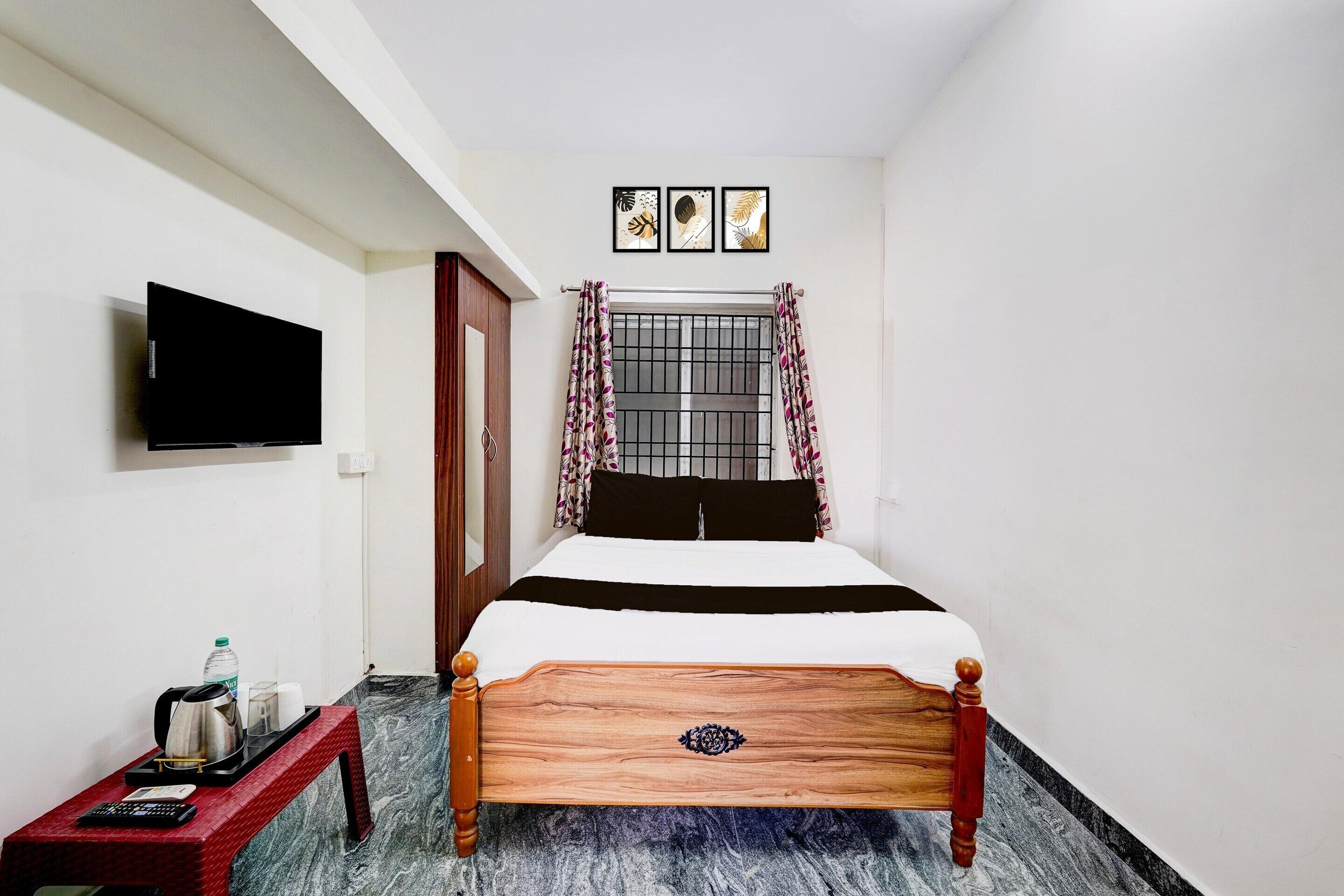 A bedroom with a bed, a TV mounted on the wall, a window with curtains, and a small table with a kettle and cups.