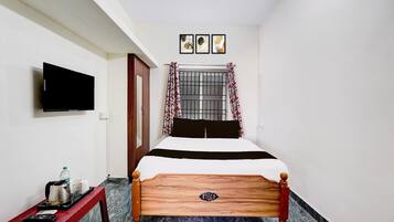 A bedroom with a bed, a TV mounted on the wall, a window with curtains, and a small table with a kettle and cups.