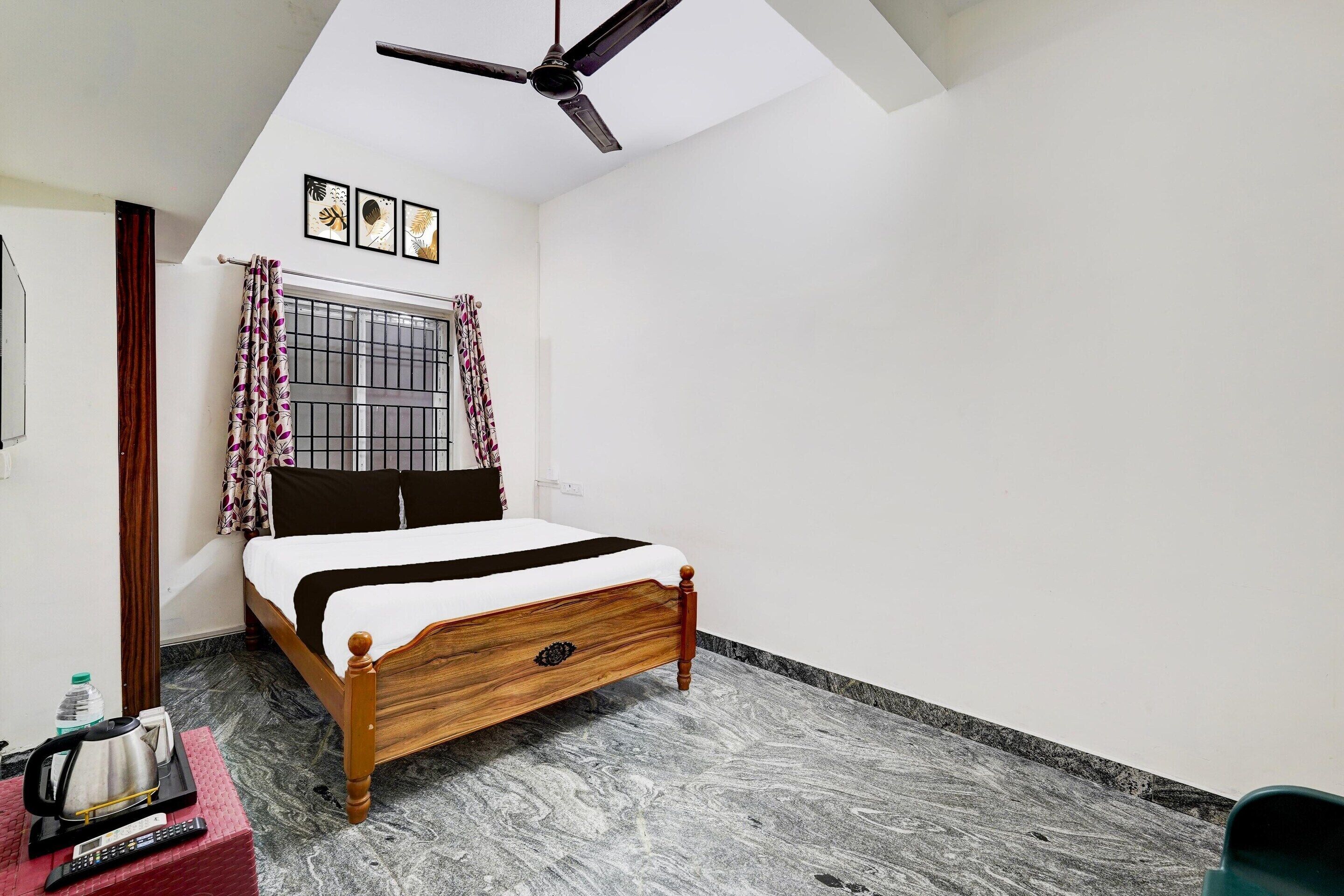 Une chambre à coucher avec un lit, un ventilateur de plafond, une fenêtre avec des rideaux et une petite table sur laquelle se trouvent une bouilloire et une télécommande.