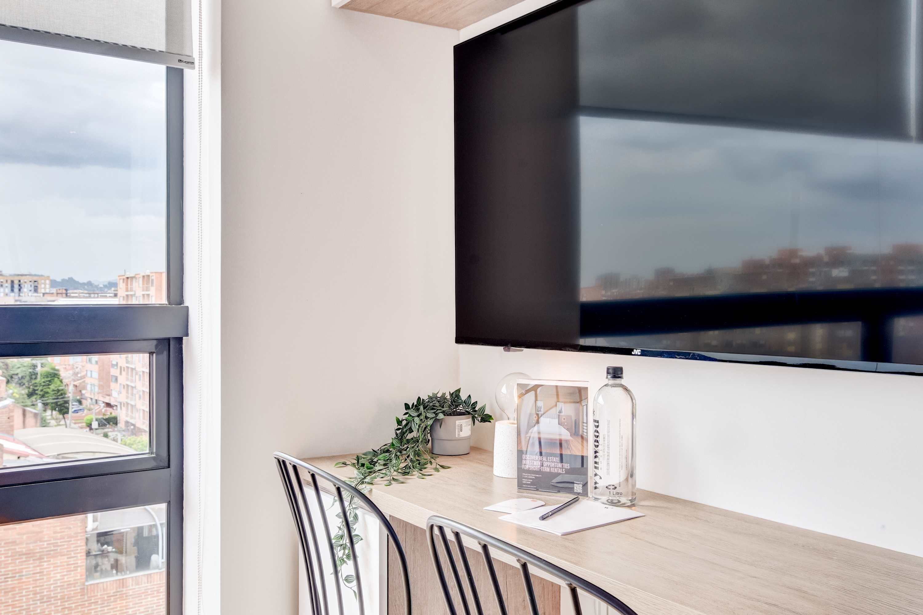 Quarto moderno com uma janela grande, TV de tela plana, uma mesinha com uma planta e uma garrafa de água.