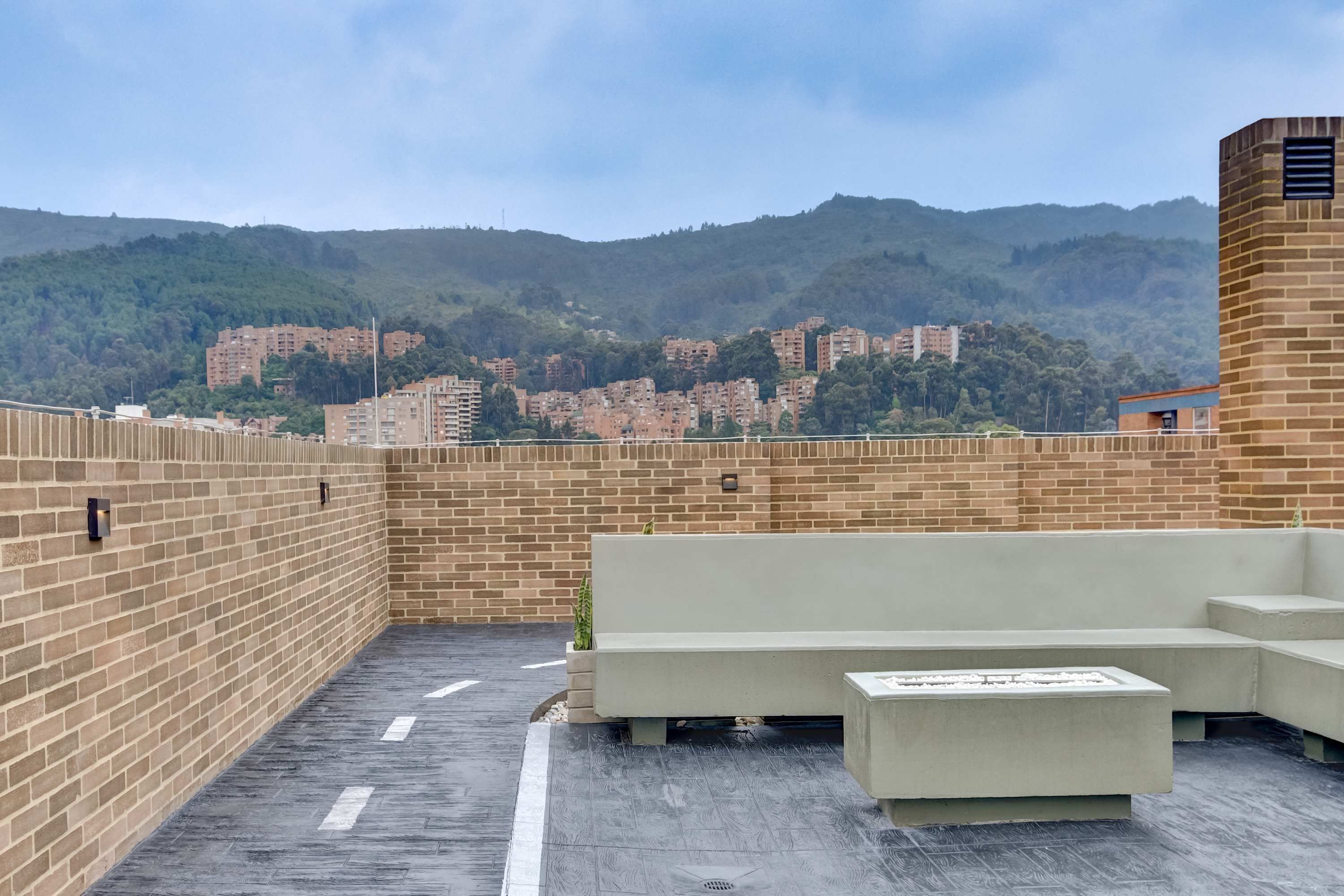 A rooftop terrace with a brick wall, a concrete bench, and a view of a cityscape with buildings and hills in the background.