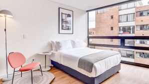Ein modernes Schlafzimmer mit großem Fenster, einem Bett, einem roten Sessel, einer Stehlampe und einem gerahmten Bild an der Wand.