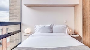 Ein modernes Hotelzimmer mit einem großen Bett, einem Nachttisch mit Lampe, Blick auf die Stadt durch ein Fenster und einer Wand mit Holzvertäfelung.