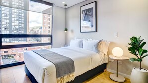 A modern bedroom with a large window, a bed with white linens, a bedside table with a lamp, and a potted plant. - w Signature Loft in the Heart of Bogot (Bogotá)