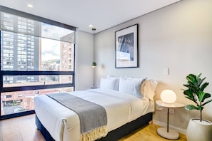 A modern bedroom with a large window, a bed with white linens, a bedside table with a lamp, and a potted plant. - w Signature Loft in the Heart of Bogot (Bogotá)