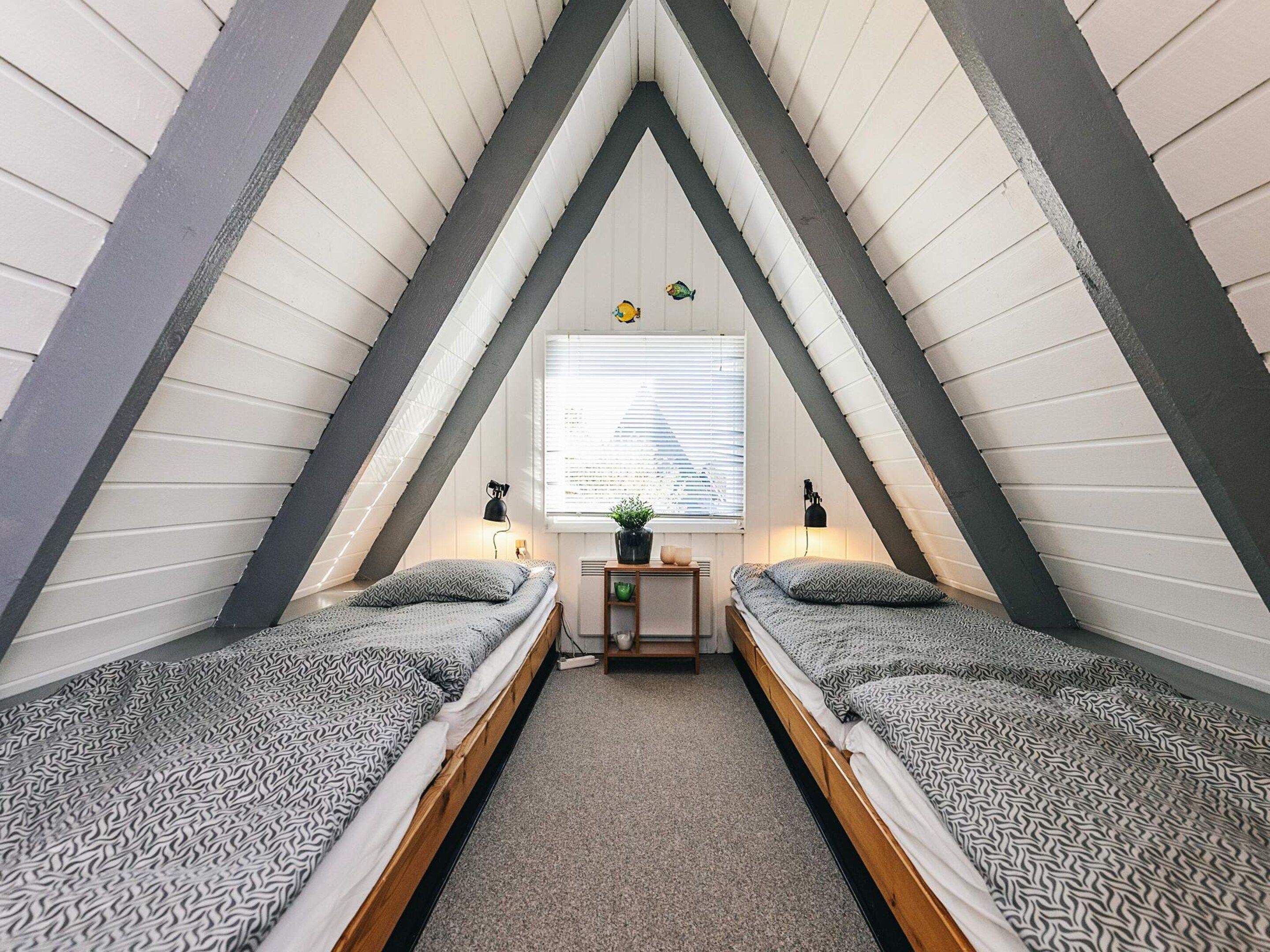 Une petite chambre mansardée triangulaire, avec deux lits, une fenêtre avec des stores, une petite table sur laquelle est posée une plante, et deux lampes fixées au mur.