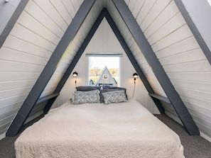 Een slaapkamer met een schuine plafond, een bed met een gestreept laken, twee wandlampen en een raam met uitzicht op een huis.