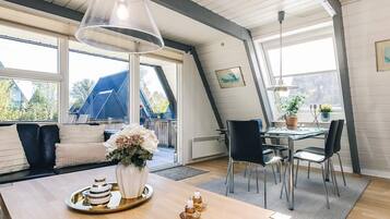 A modern interior with a dining area, a sofa with cushions, a table with a lamp, and a view of the outside.