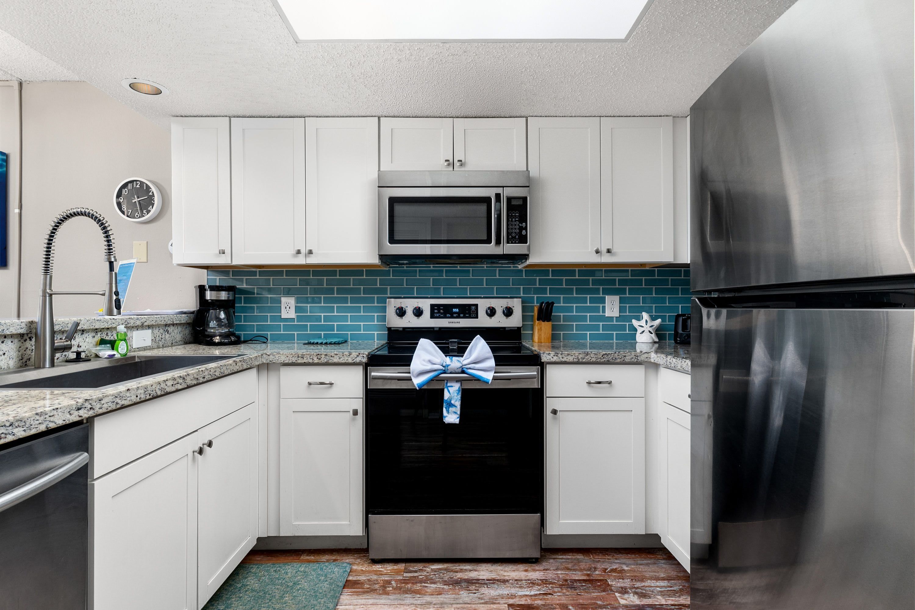Una cocina con gabinetes blancos, microondas, horno y refrigerador de acero inoxidable, salpicadero azul turquesa y un reloj en la pared.