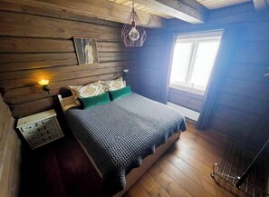 Un dormitorio rústico tipo cabaña de madera, con cama, mesita de noche, lámpara de pared y ventana.