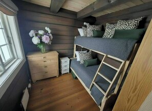 A bunk bed with patterned pillows, a wooden dresser, a small white cabinet, and a vase of flowers.