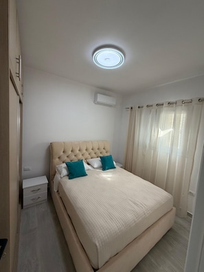 Une chambre à coucher comprenant un lit, une table de chevet, un appareil de climatisation et une fenêtre avec des rideaux.