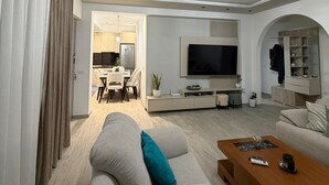 A modern living room with a sofa, coffee table, TV, and a dining area in the background. - Cozy Family Villa close to the beach! (Vlora)