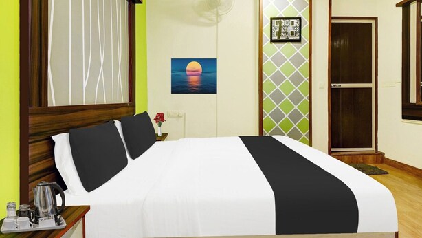 A hotel room with a bed, a nightstand, a picture of a sunset, a fan, and a door. - Deluxe GL Classic (Alto de Porvorim)
