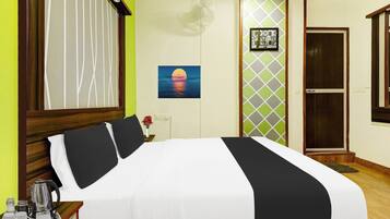 A hotel room with a bed, a nightstand, a picture of a sunset, a fan, and a door.