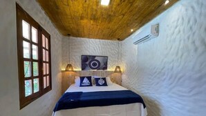 Un dormitorio con techo de madera, una cama con cubrecama blanco, dos lámparas fijadas a la pared y un ventanal con marcos de madera.