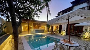 Una piscina con cubierta de madera, mobiliario exterior y un edificio con techo de paja.