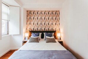 Ein Hotelzimmer mit einem großen Bett, zwei Nachttischen mit Lampen, einem gemusterten Kopfteil und einem Fenster links.