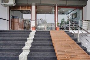 Un escalier menant à l’entrée d’un bâtiment, avec une rampe en bois sur la droite, des portes en verre et un ventilateur de plafond.