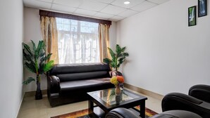 Une pièce comprenant un canapé en cuir noir, une table basse en verre, deux plantes en pot, une fenêtre avec des rideaux et un cadre photo fixé au mur.