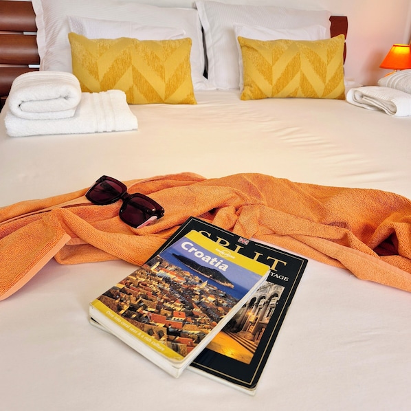 A neatly made bed with white linens, two yellow pillows, a folded white towel, a pair of sunglasses, an orange blanket, and a travel guide book titled 'Croatia' on the bed.
