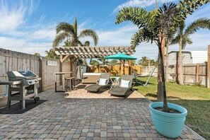 Un patio trasero con una parrilla, sillas de descanso, un pérgola y un árbol en maceta.